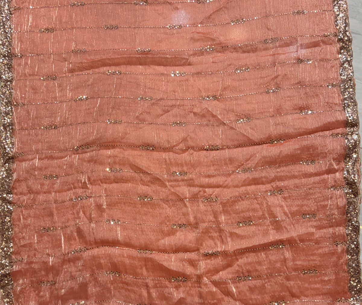 A square peach-colored fabric scarf or shawl with metallic gold embroidered details and a gold border, laid flat on a gray tiled floor.