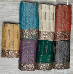 Six folded pieces of embellished fabric in different colors are arranged on a gray floor. The fabrics include gold, teal, off-white, rust red, purple, emerald green, charcoal gray, and navy blue, each featuring vertical sequin patterns and a wide sequined border.