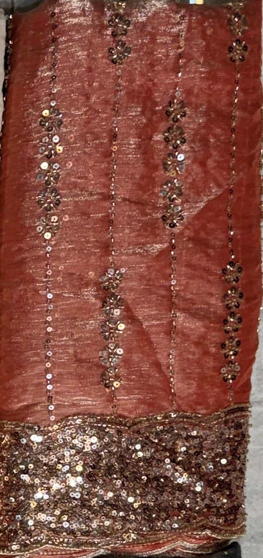 A close-up of a rust-red or terracotta-colored fabric with vertical rows of metallic sequins and a heavily embellished sequined border along the bottom.