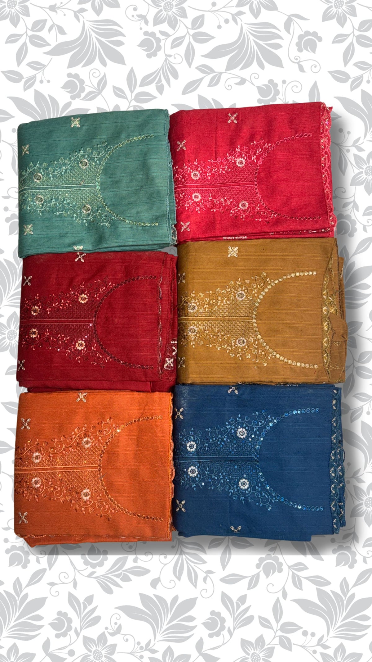 Six folded cotton fabric pieces in teal, red, maroon, mustard, orange, and navy blue, each with white embroidered floral and geometric patterns, arranged in a 2x3 grid on a white background with a gray floral pattern.