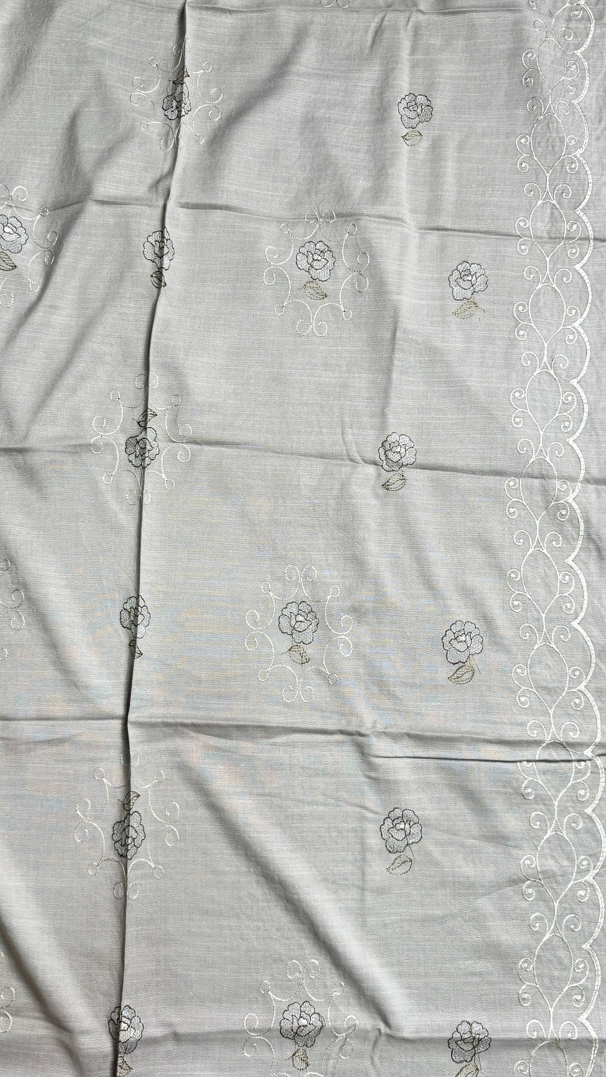 A close-up of a light gray fabric with delicate white floral embroidery and a white scalloped lace border along the right edge.
