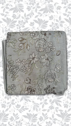 A folded piece of light gray fabric with intricate silver and gold floral embroidery, displayed on a white background with a gray floral pattern.