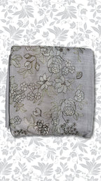 A folded light gray fabric piece with an all-over floral embroidery pattern in gray, black, and subtle gold thread, set against a white background with a gray floral border.