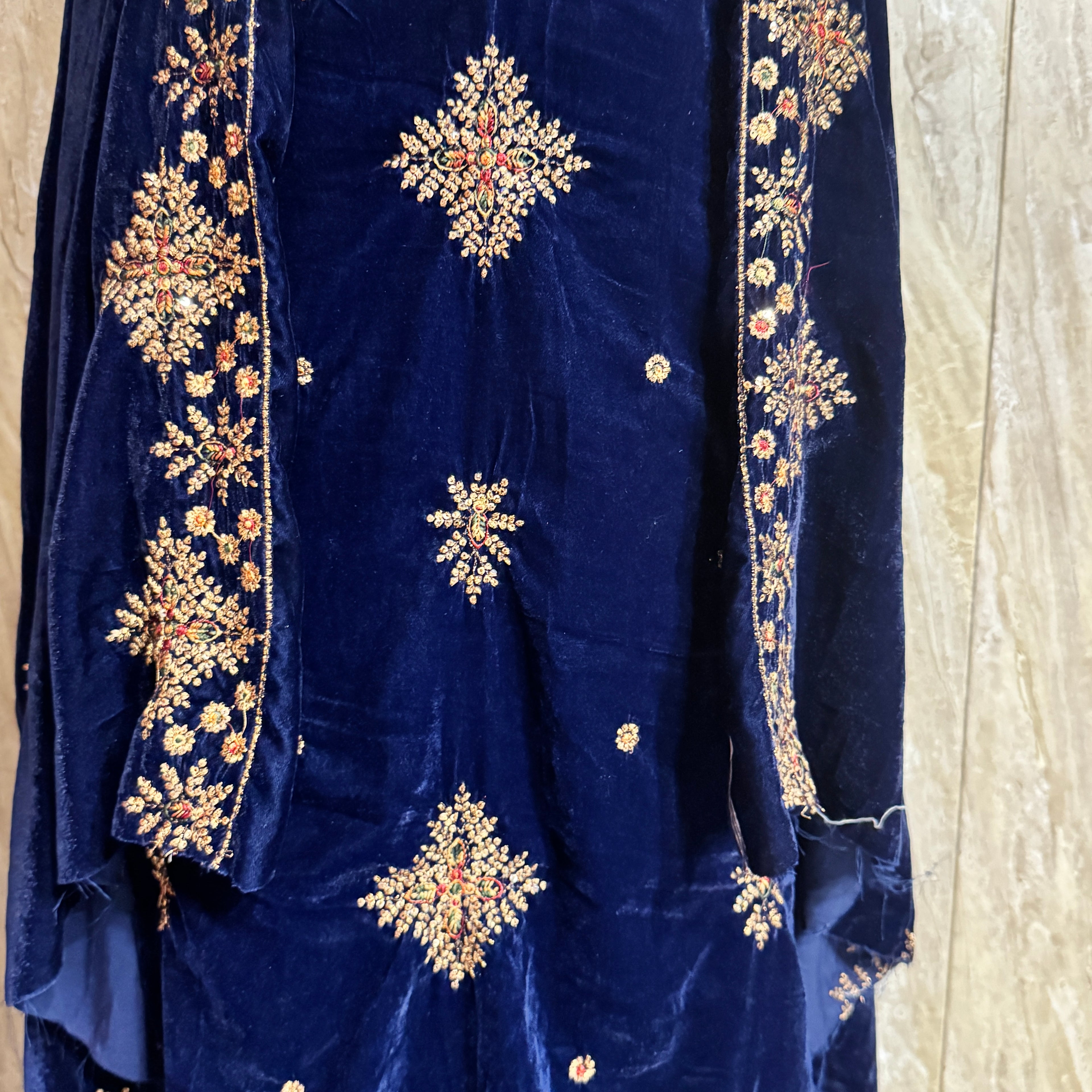 A close-up of a navy blue velvet garment, likely a traditional tunic or dress, featuring intricate gold, red, and green embroidered patterns on the front and sleeve borders.