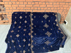 A navy blue velvet throw blanket with gold embroidered snowflake and star patterns draped over a gray sofa in front of a brick wall.