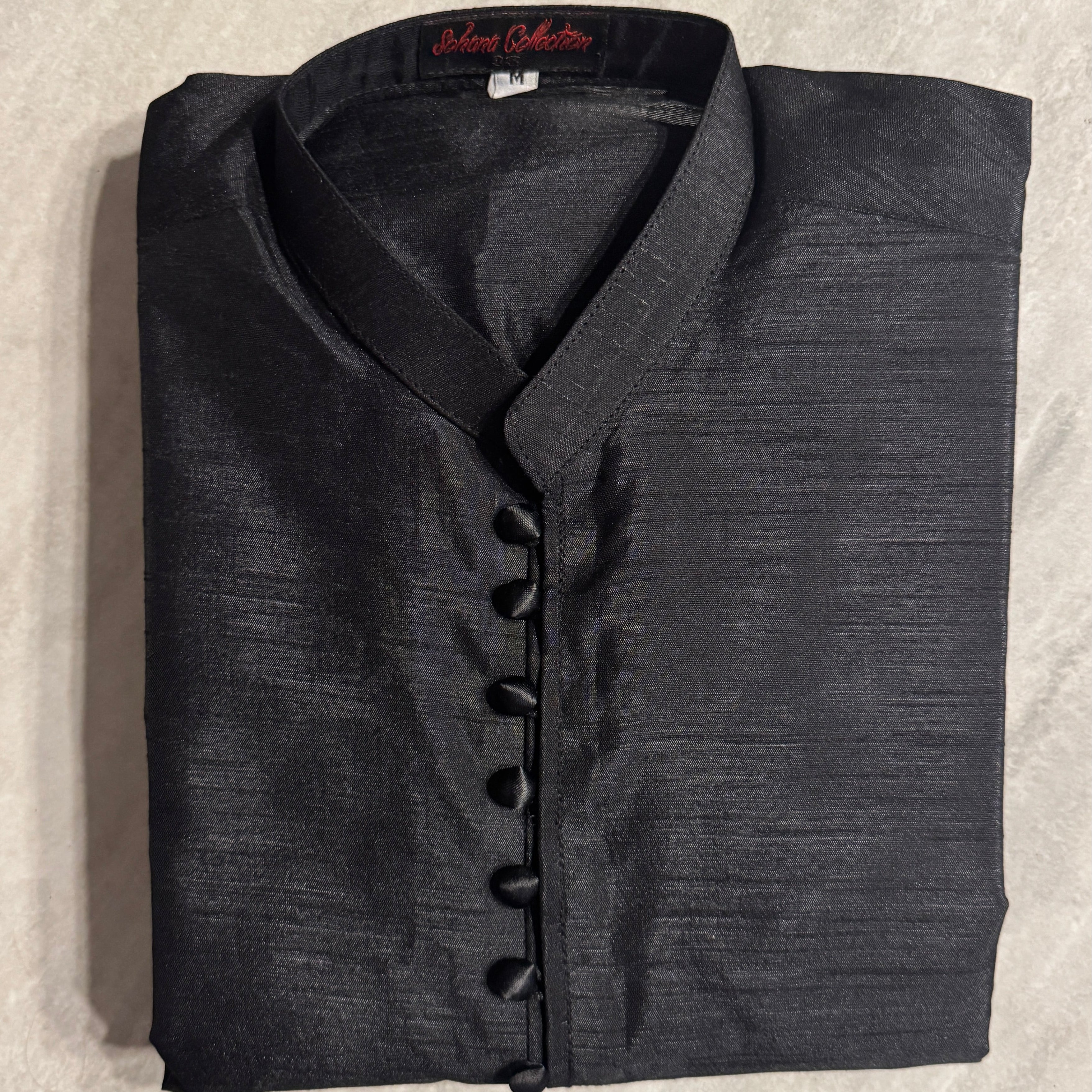 A folded black textured traditional shirt with a mandarin collar and button placket, laid flat on a light gray tiled surface. The brand label inside the collar reads 'Delhi Outfitter'.