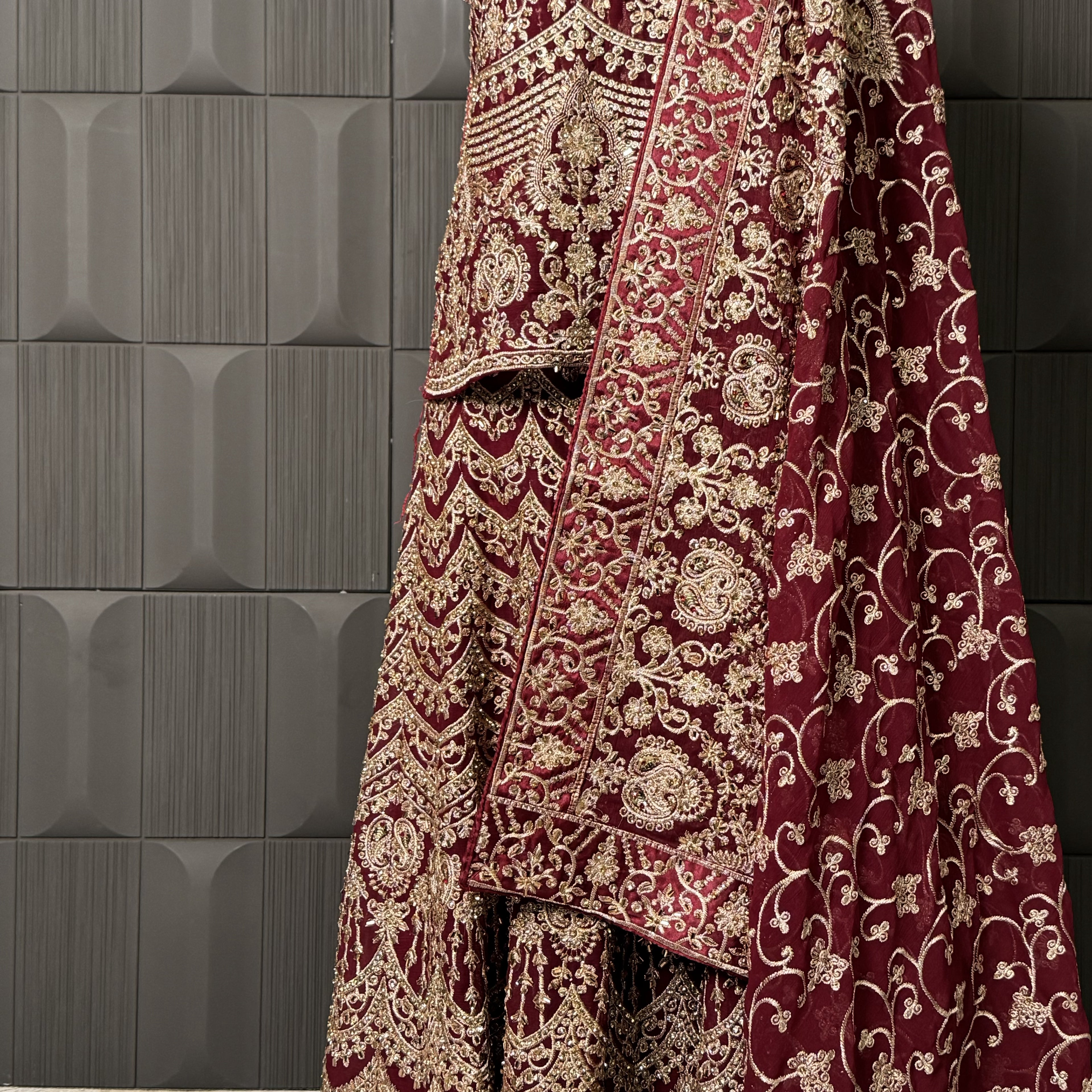 Maroon embroidered fabric with gold patterns against a dark tiled wall INDIAN LEHENGA MACHINE WORK
