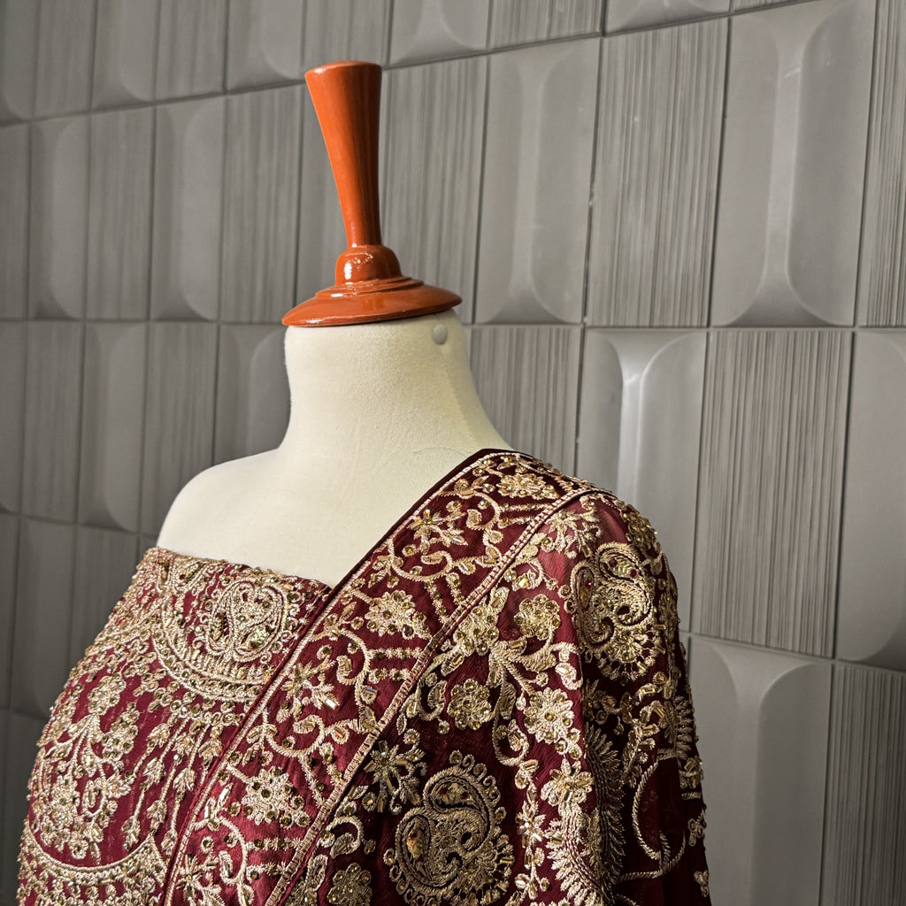 Mannequin wearing a maroon saree with gold embroidery against a gray tiled wall.