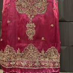 A close-up of a magenta pink fabric garment featuring intricate gold zari embroidery, floral motifs, and a heavily embellished border along the bottom hem.