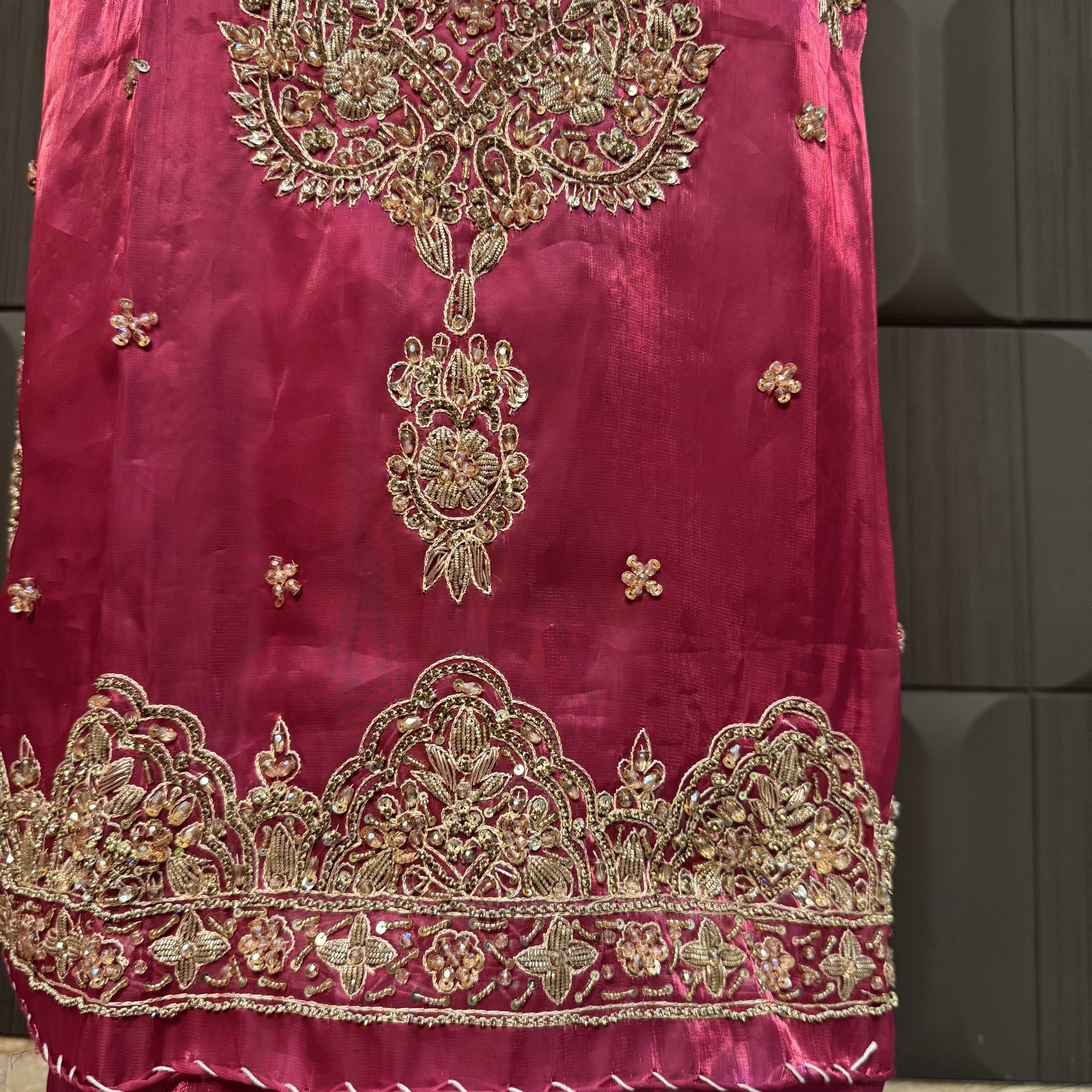 A close-up of a magenta pink fabric garment featuring intricate gold zari embroidery, floral motifs, and a heavily embellished border along the bottom hem.