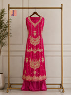 A bright pink traditional dress with intricate gold embroidery displayed on a wooden hanger hanging from a gold clothing rack, set against a light beige paneled wall.