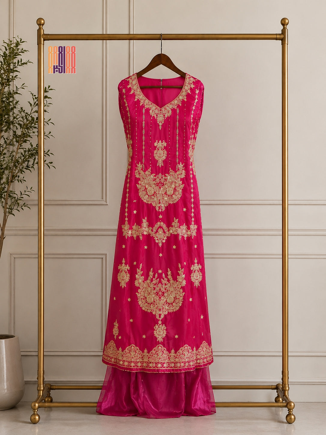 A bright pink traditional dress with intricate gold embroidery displayed on a wooden hanger hanging from a gold clothing rack, set against a light beige paneled wall.