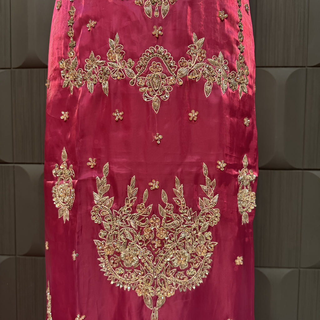 A close-up of a magenta pink fabric garment featuring intricate gold and silver zari embroidery with floral and paisley patterns, displayed against a dark gray textured background.