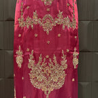 A close-up of a magenta pink fabric garment featuring intricate gold and silver zari embroidery with floral and paisley patterns, displayed against a dark gray textured background.