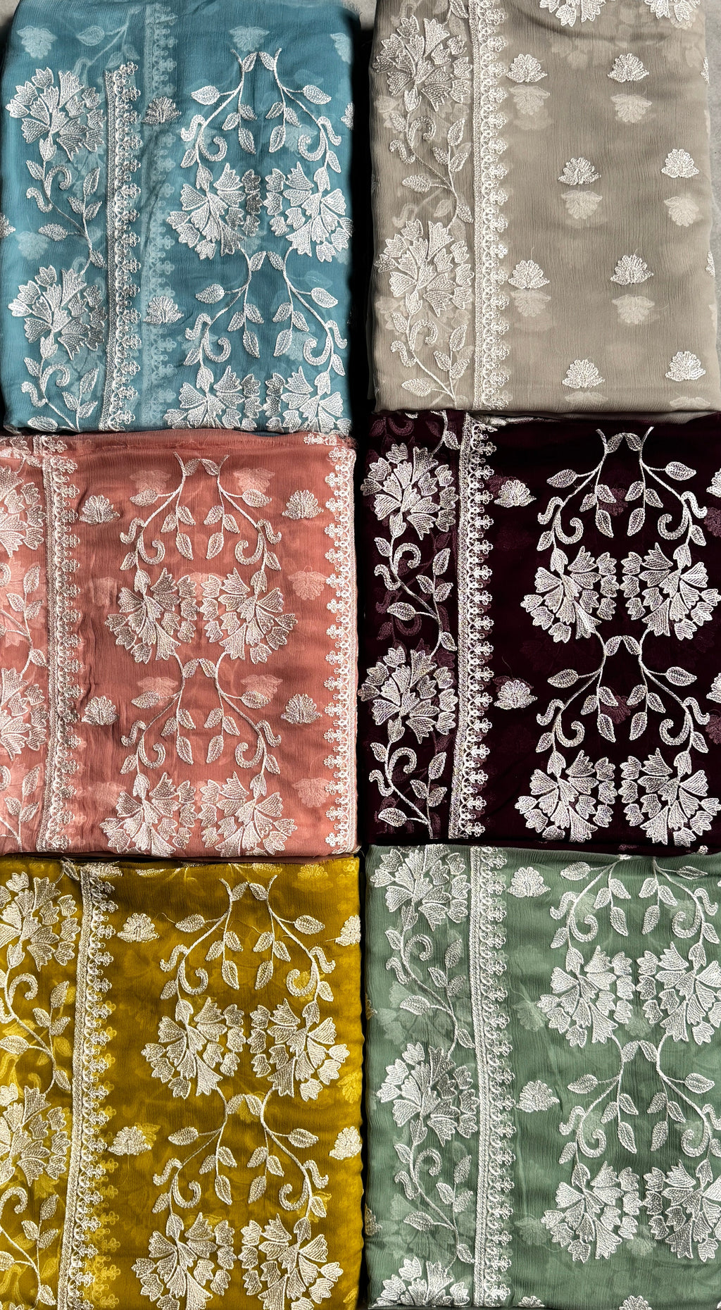 Six folded fabric pieces in different colors arranged in a 2x3 grid on a gray floor. The fabrics feature intricate white floral embroidery and border detailing.
