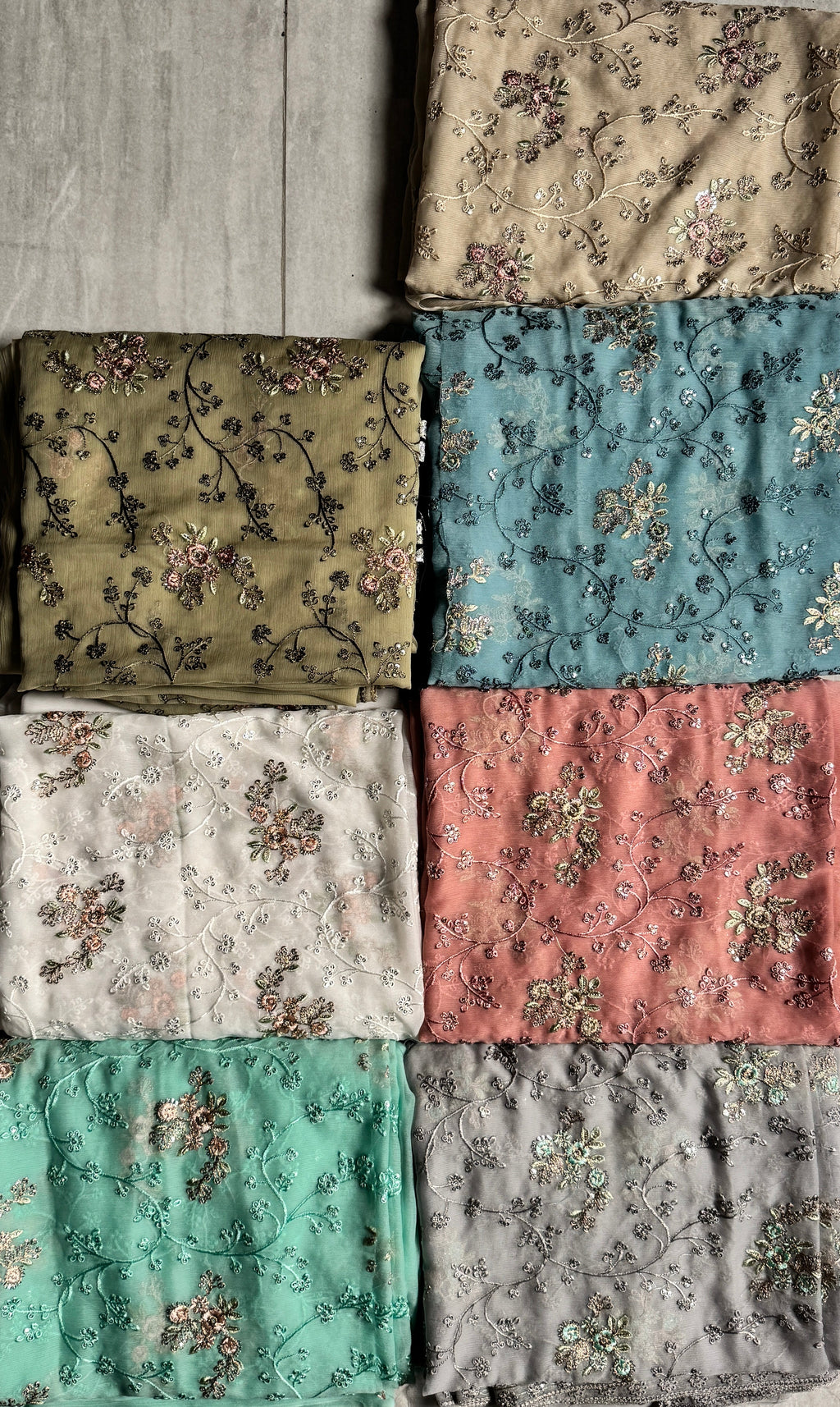 Eight folded fabrics with floral patterns in various colors, arranged in a grid on a gray wood floor.