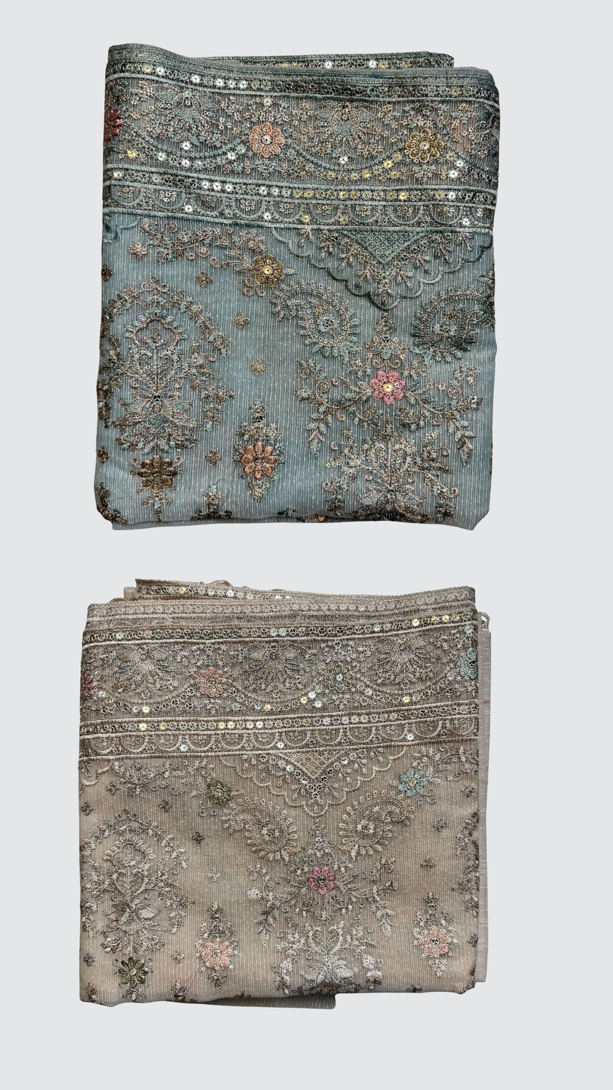 Two folded pieces of ornate embroidered fabric shown vertically on a light gray background. The top piece is dusty blue with silver mirror work and floral embroidery, while the bottom piece is a warm taupe-beige with similar silver embellishment.