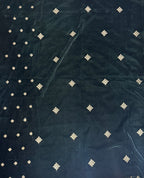 A dark green velvet fabric, likely a saree pallu or dupatta, laid flat on a light-colored tiled floor. The fabric features a border of light green along the edges and is decorated with rows of small, metallic gold diamond-shaped motifs.