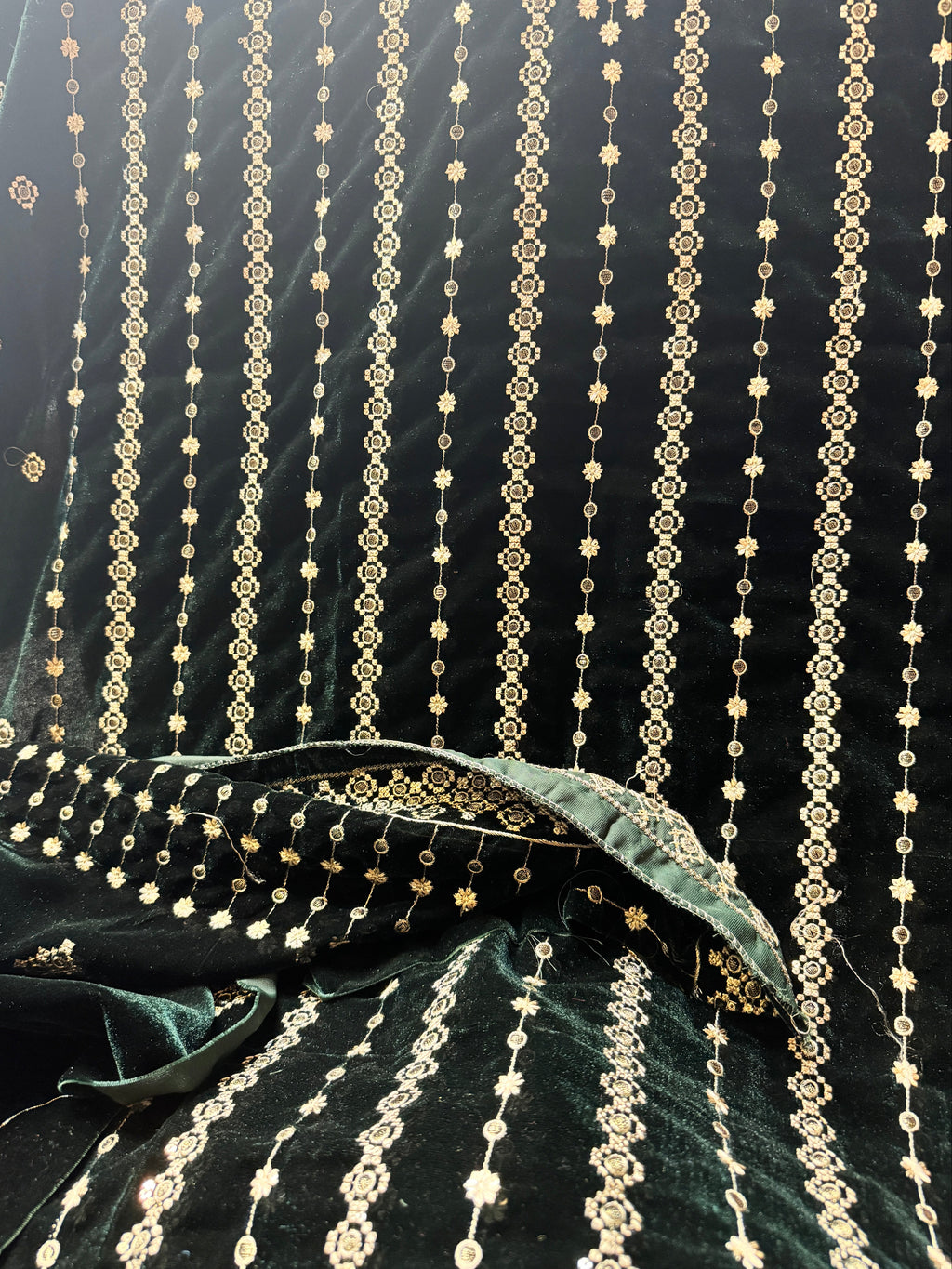 Dark green velvet fabric draped to show its texture and intricate gold zari embroidery, featuring vertical strands of metallic gold beadwork and floral motifs.