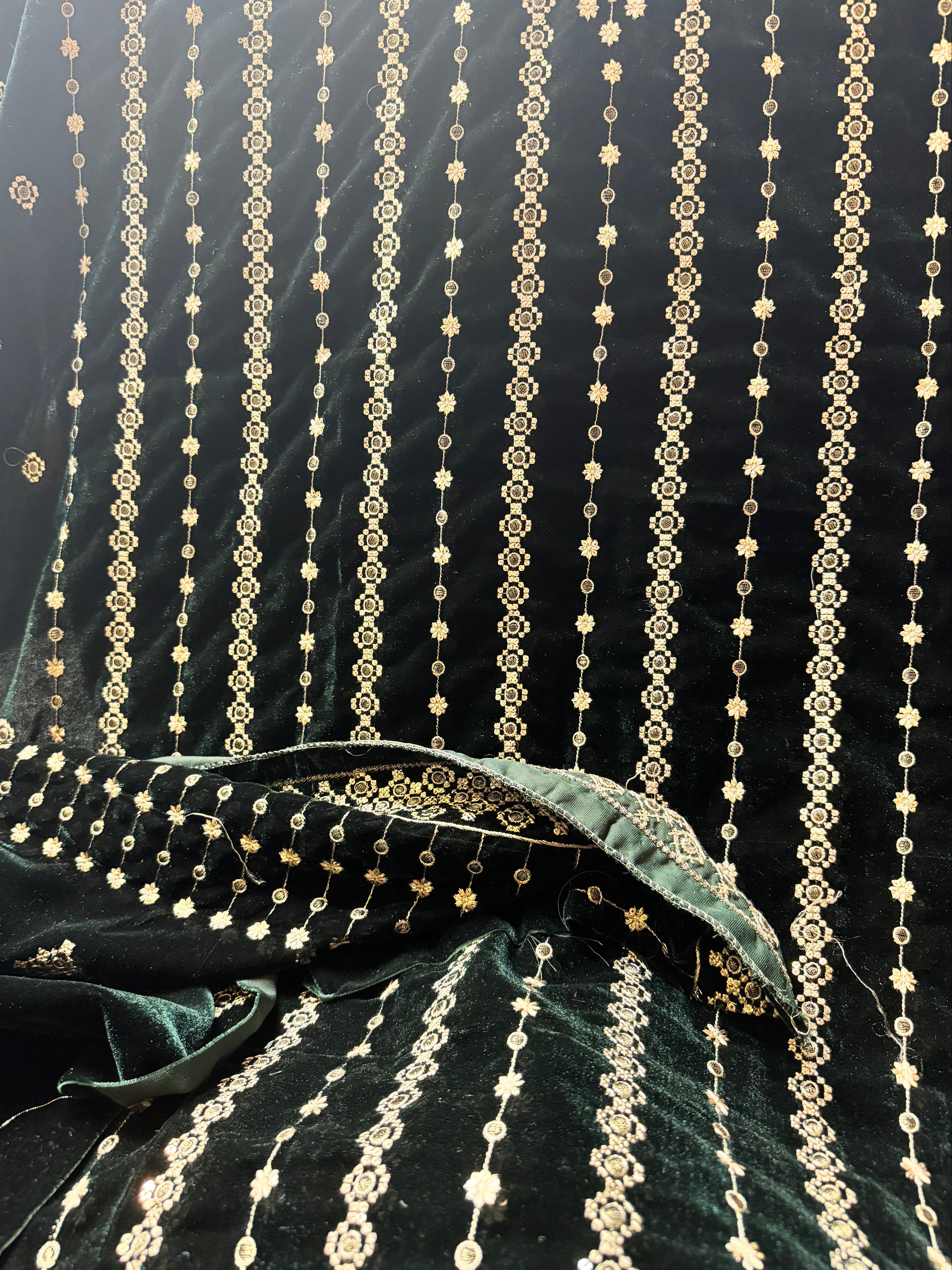 Dark green velvet fabric draped to show its texture and intricate gold zari embroidery, featuring vertical strands of metallic gold beadwork and floral motifs.